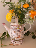 Cream ceramic watering can with hand-painted terracotta, teal, and ochre florals, bird motif on body, mauve-tipped spout.