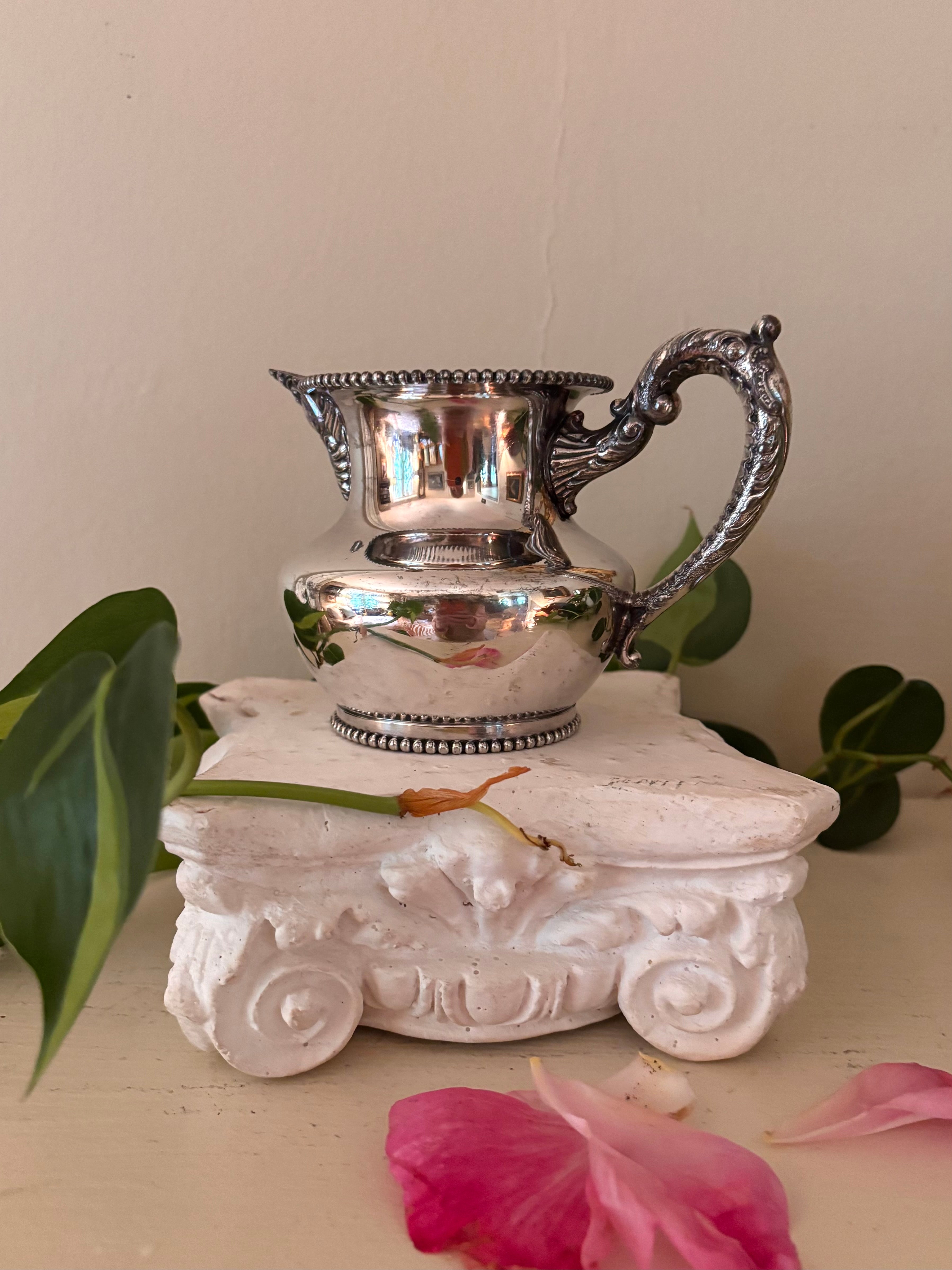 Silver-plate cream pitcher showing ornate acanthus-flanked pour spout and beaded borders at rim and foot.