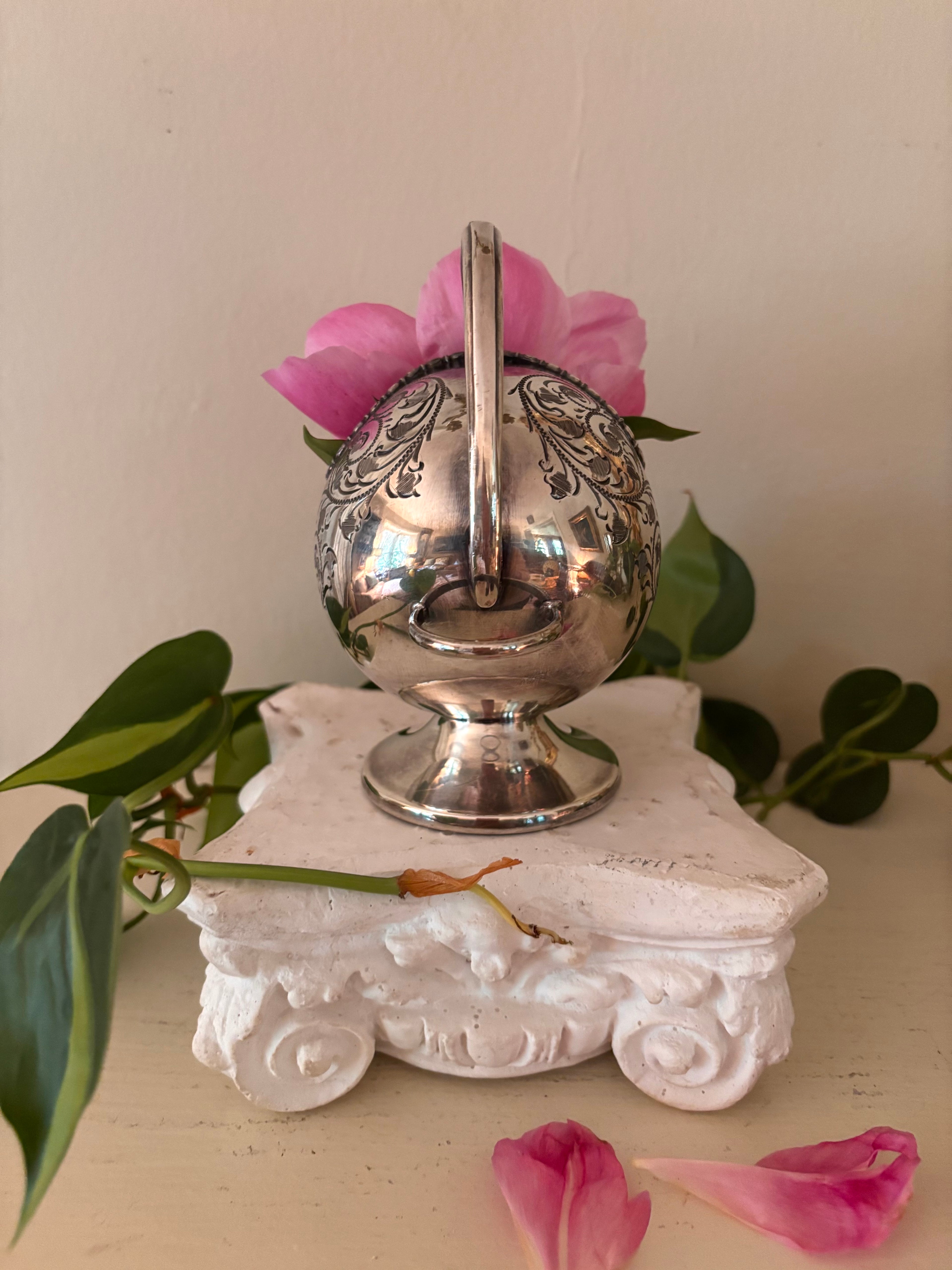 Rear view of silver-plate sugar scuttle showing high arching handle, ball finial, and scroll-engraved round body.