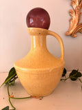 Speckled golden yellow ceramic jug with single handle and deep plum glass orb resting in neck.