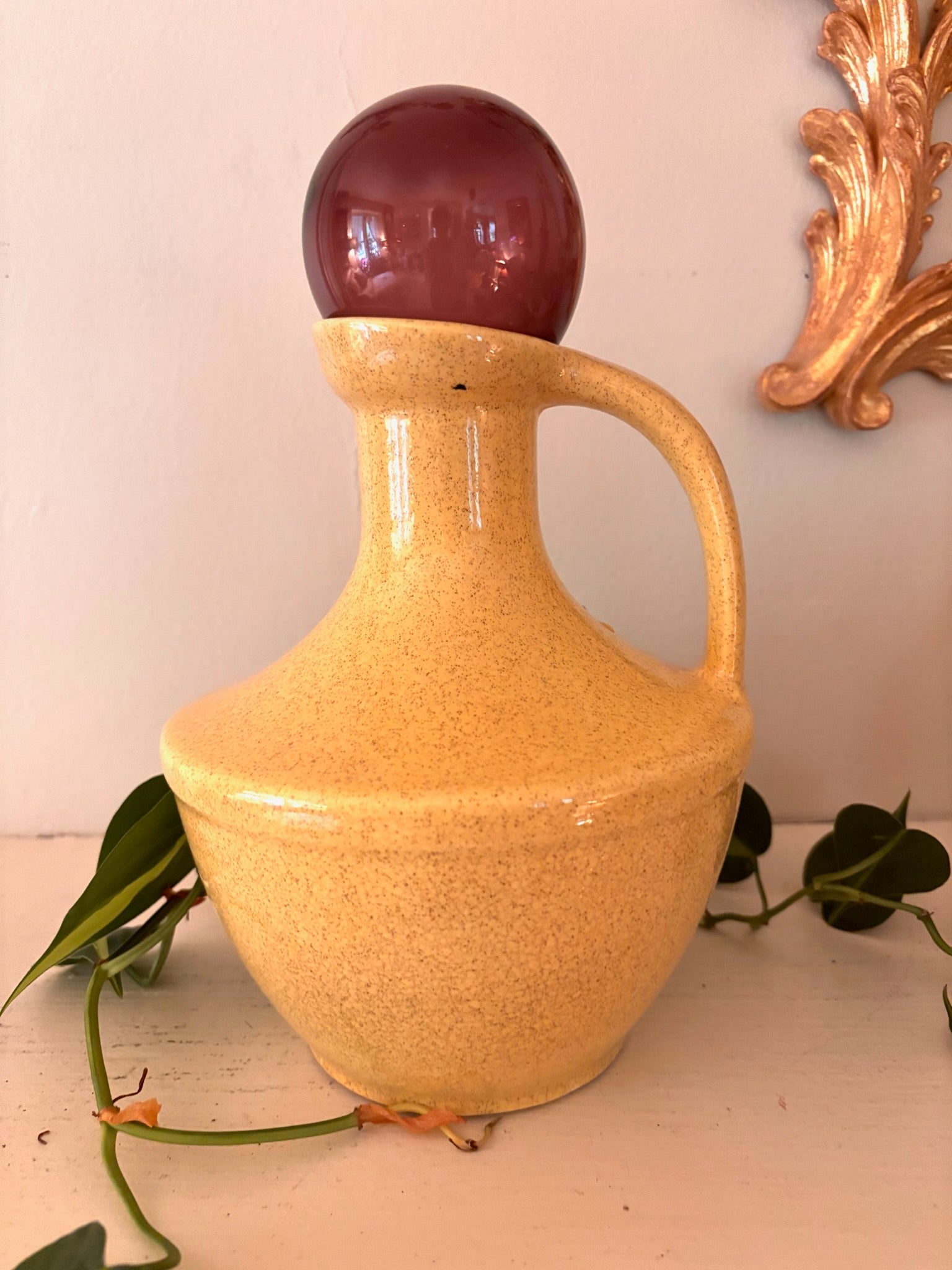 Speckled golden yellow ceramic jug with single handle and deep plum glass orb resting in neck.