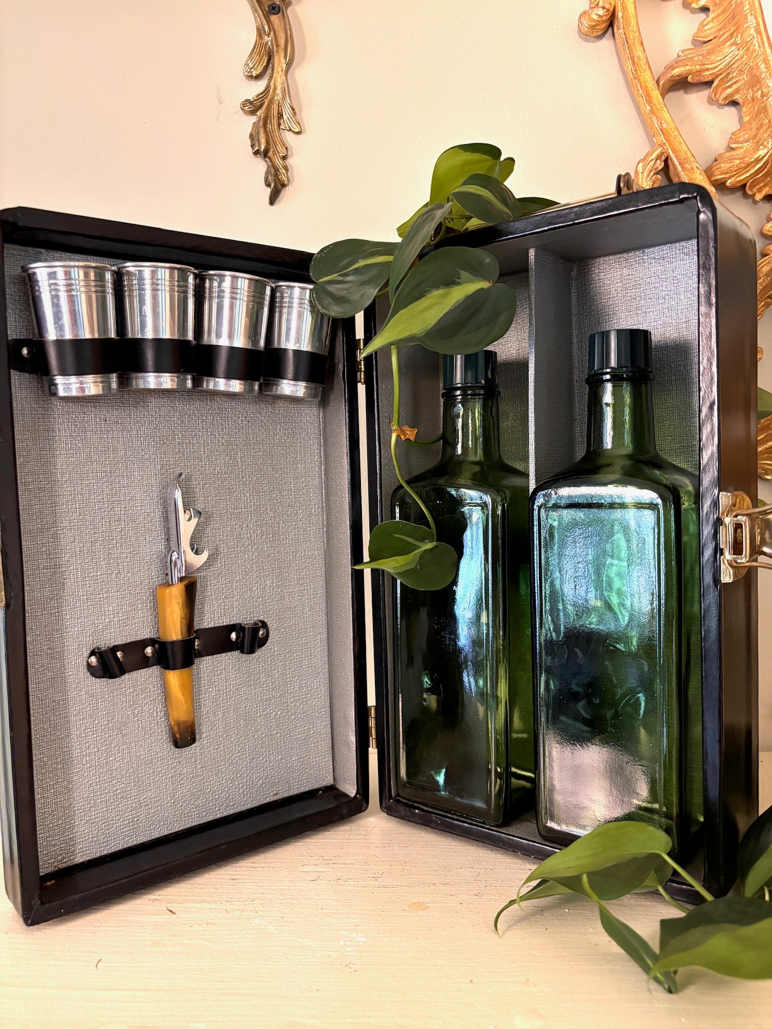 Open travel bar case showing chrome cups, amber-handled corkscrew, and two square green glass bottles.