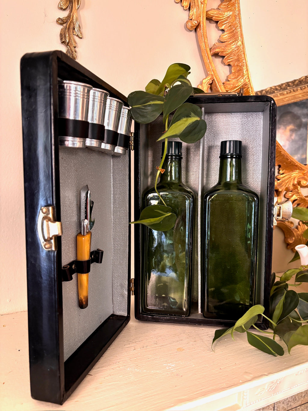 Open black travel bar case with four chrome cups, an amber-handled opener, and two green glass bottles inside.