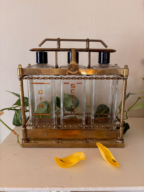 Antique brass tantalus liquor caddy with three glass decanters stenciled in gold with spirit names including Gin and Scotch, with twisted rope rail detailing, turned corner columns, and Bakelite stoppers