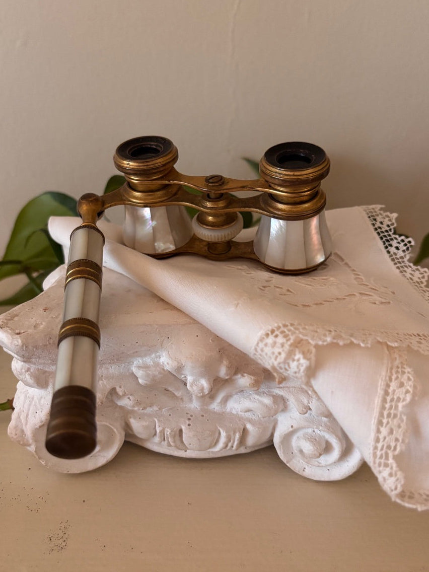Antique opera glasses with iridescent mother of pearl bodies and brass hardware, with an extending mother of pearl and brass handle, resting on a white plaster architectural fragment with a linen handkerchief.