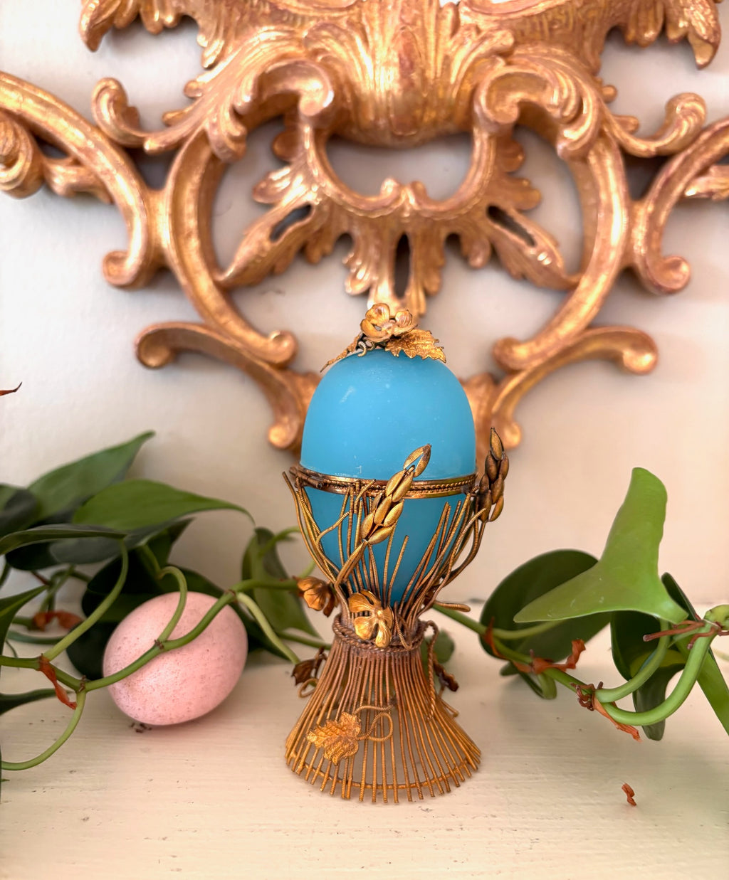 Sky blue opaline glass egg held in an ornate gilt metal wheat-sheaf stand with leaves and a floral top.