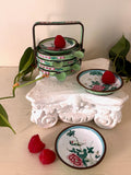 Stacked Canton enamel dishes in a green enamel handled basket beside a painted enamel dish with peonies.