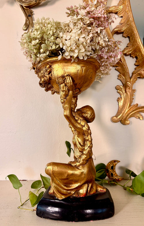 Side view of a gilded planter stand, hand supporting the floral bowl above the figure's head.