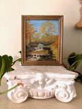 Framed oil painting of a stream falling over rocks through gold and orange autumn woodland under a blue sky.