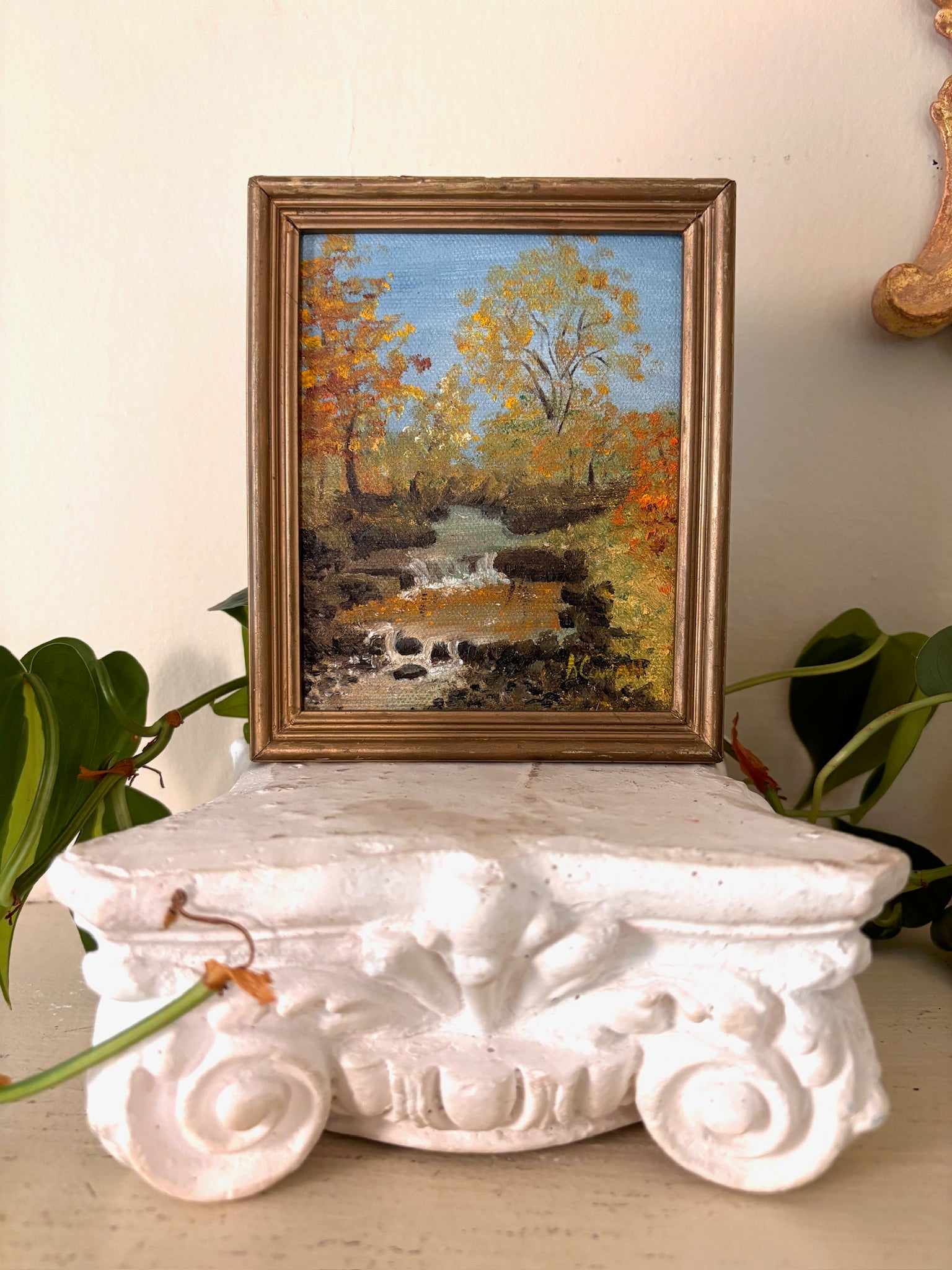 Framed oil painting of a stream falling over rocks through gold and orange autumn woodland under a blue sky.