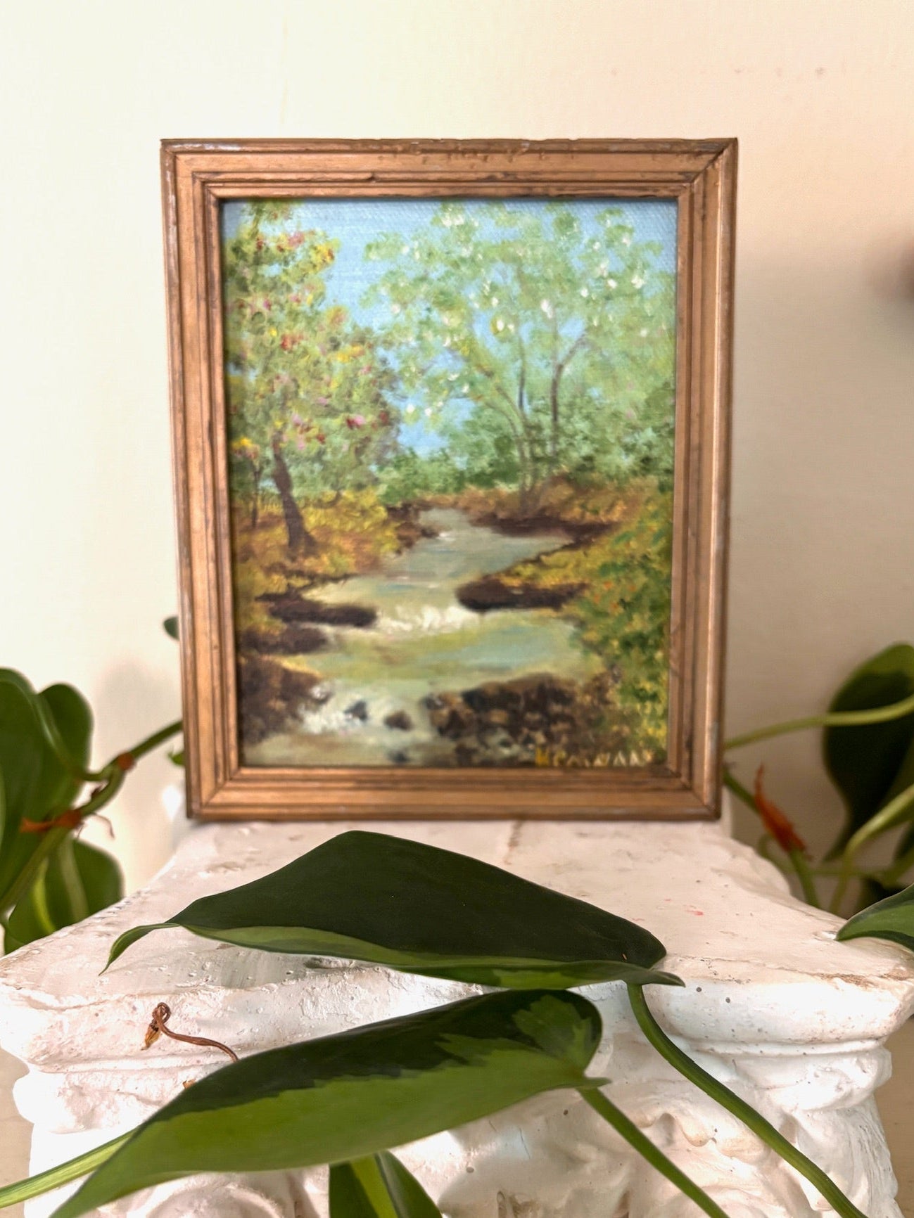 Framed oil painting of a stream winding through green woodland with autumn touches, in a carved giltwood frame.