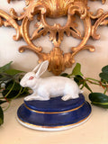 White glazed porcelain rabbit with a red eye, resting on an oval cobalt blue base with gold line trim.