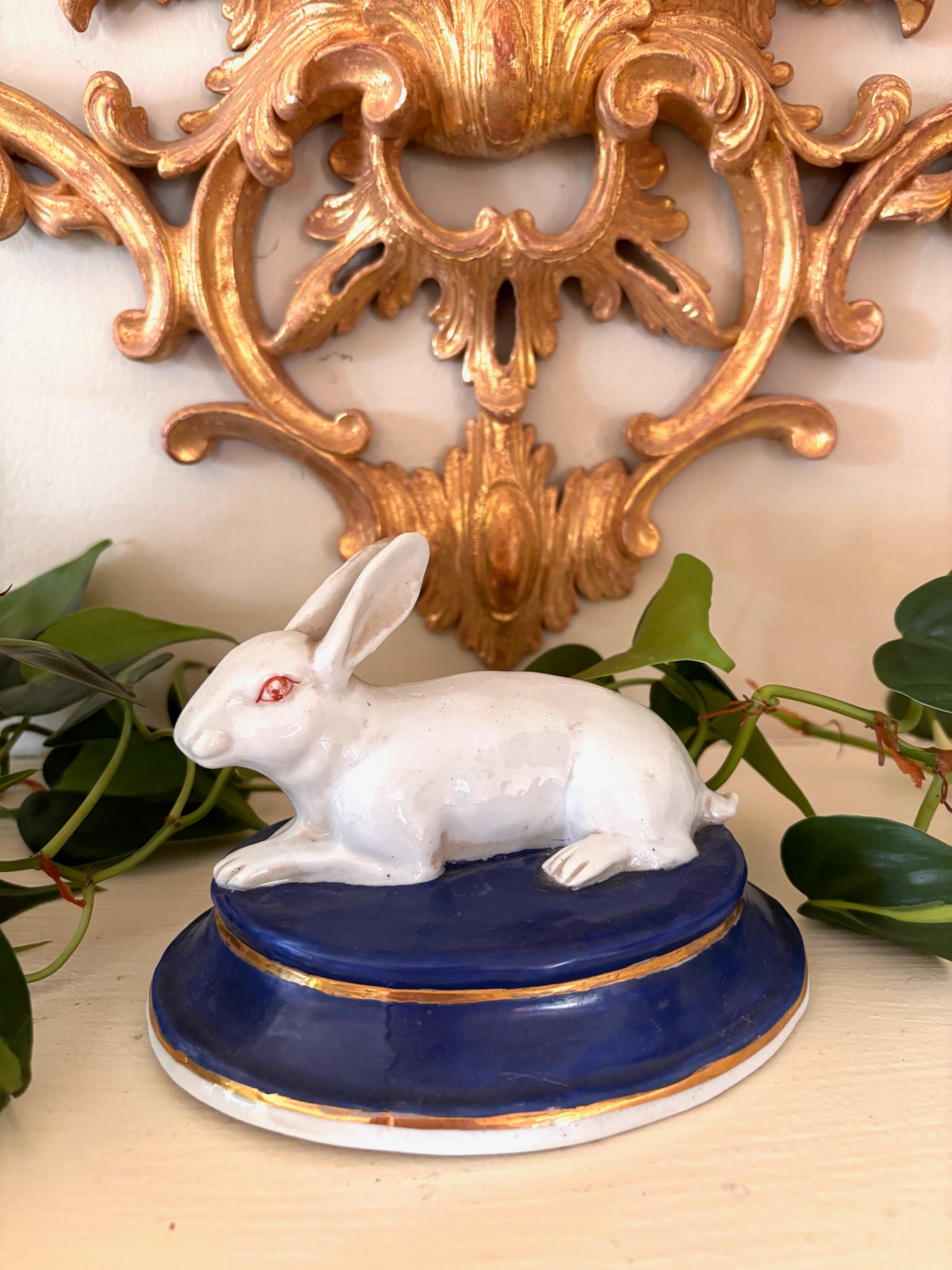 White glazed porcelain rabbit with a red eye, resting on an oval cobalt blue base with gold line trim.