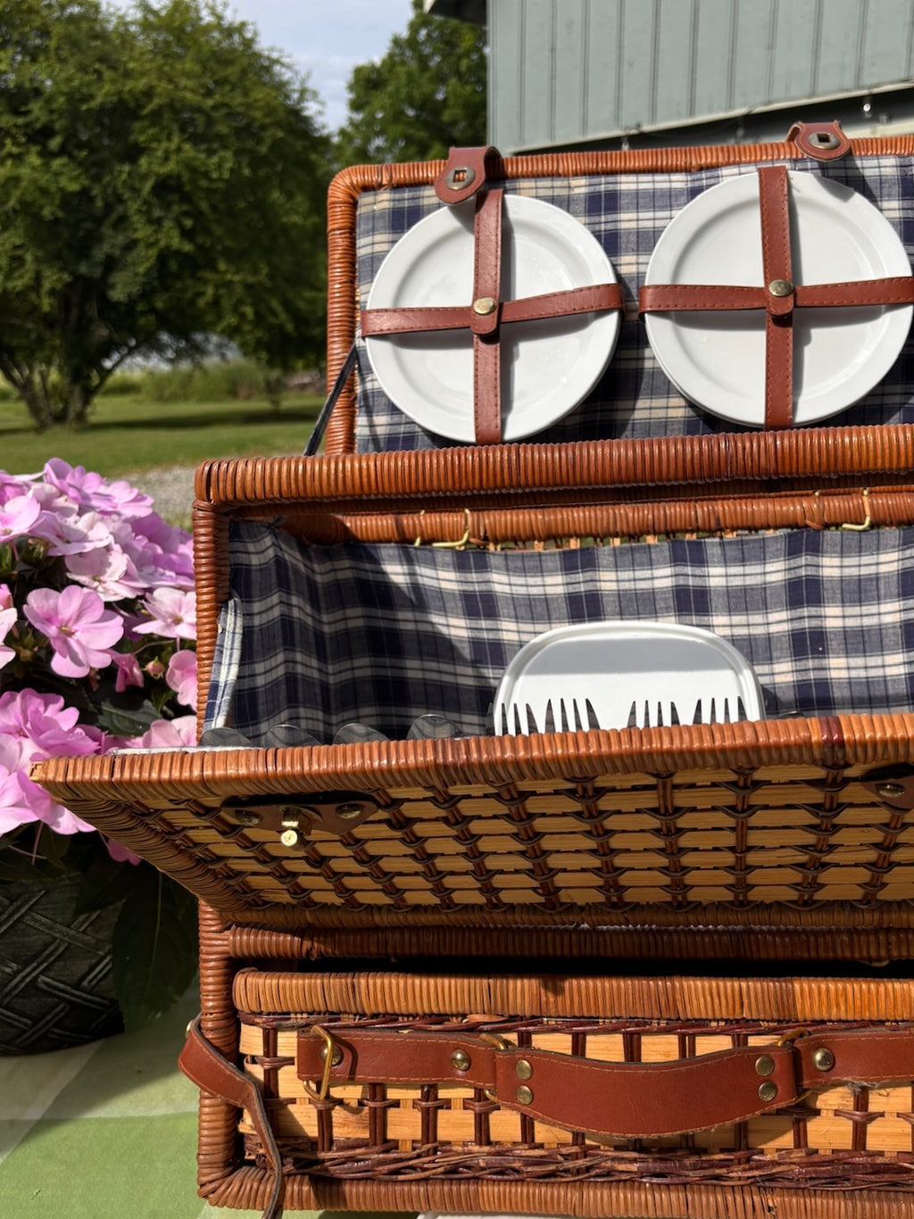 Open wicker picnic basket lid with two white ceramic plates secured by leather cross-straps.
